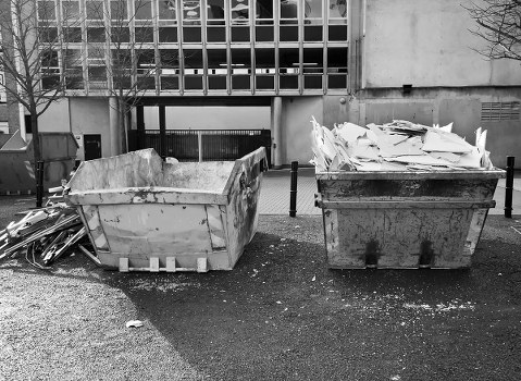 Two large industrial skips positioned side by side on a paved outdoor area in front of a modern building with a glass and concrete facade. The left skip contains broken wood and debris with rough, splintered edges and a weathered, slightly rusted surface. The right skip is filled with crumpled, torn paper and miscellaneous waste materials, appearing overfilled with some waste spilling slightly over the top edge. The ground beneath the skips is covered with small scattered fragments and dust, indicating recent use. In the background, part of a building's metal railing and entrance are visible, along with some vertical posts, suggesting this area may be used for waste collection or clearance activities typical of a residential or commercial property located near Dulwich. The overall scene depicts typical rubbish removal equipment used by Dulwich House Clearance in the Dulwich area of London, emphasizing the practical aspect of waste disposal services in the local context.