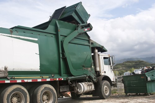 A large green skip bin mounted on the chassis of a white refuse collection truck parked on a gravel driveway with a backdrop of cloudy sky and distant hills. The skip, made of metal with a textured, weathered surface, is partially raised and filled with mixed waste materials, including black rubbish bags and assorted debris. The truck features a side mirror and visible exhaust pipe, with the cab situated on the right side of the image. Nearby, a green wheeled waste container is positioned to the right of the truck, also filled with rubbish. The setting appears to be an outdoor area suitable for waste collection, with natural light illuminating the scene. The scene aligns with services offered by Dulwich House Clearance, specialising in rubbish removal and house clearance in Dulwich and surrounding areas, including postcode regions near London.