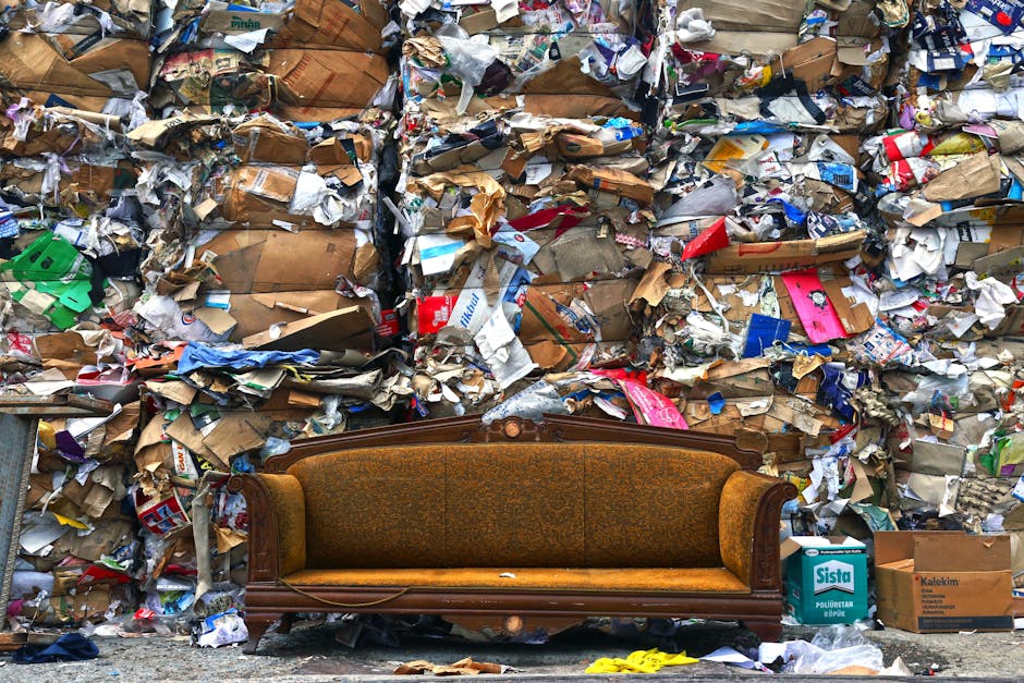 A vintage brown and orange upholstered sofa with ornate wooden trim on the arms and backrest is positioned in front of a large, dense wall of tightly stacked cardboard and paper waste. The waste inclu